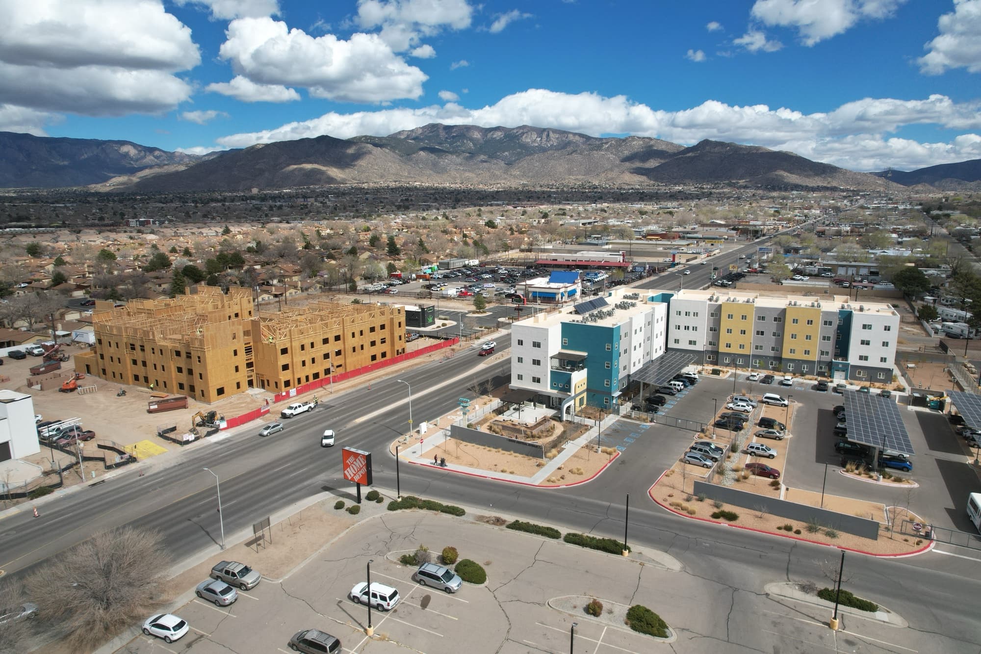 Aerial photograph of a city street with a large wooden building under construction on the left, and a completed multi-colored apartment building on the right. Mountains are visible in the distance.